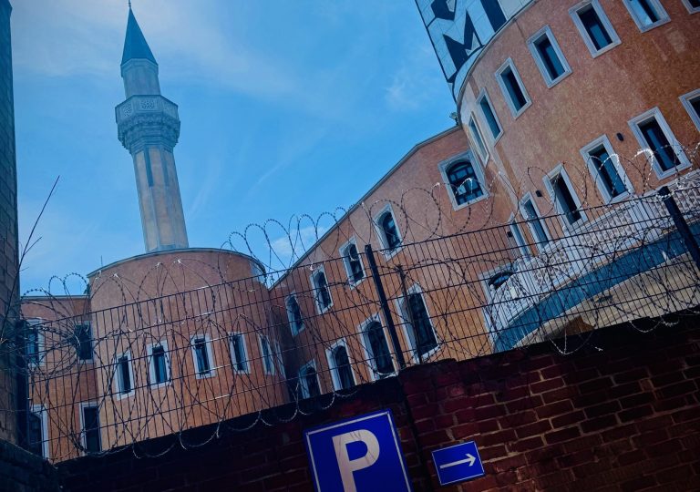 Ein Moschee-Minarett erhebt sich hinter einem Stacheldrahtzaun und einem Parkplatzschild gegen den blauen Himmel in Berlins pulsierendem Bezirk Kreuzberg.