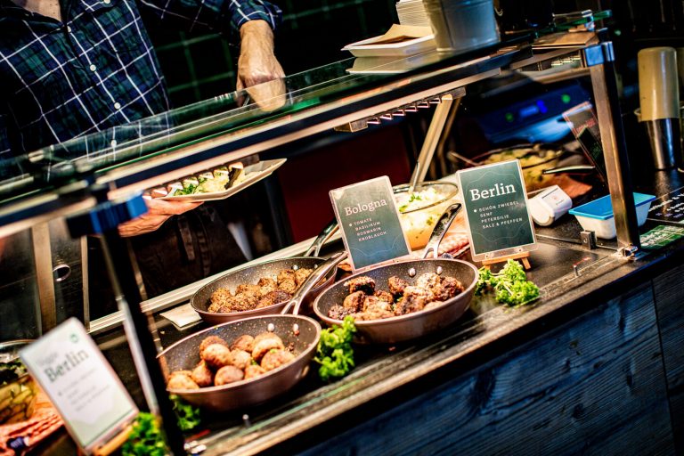 Eine Person serviert Speisen an einem Buffet mit Schildern mit der Aufschrift "Bologna" und "Berlin" und bietet so einen Vorgeschmack auf die verschiedenen Geschmacksrichtungen Kreuzbergs - wie bei einer kulinarischen Stadtführung durch die lebendigen Bezirke der Stadt.