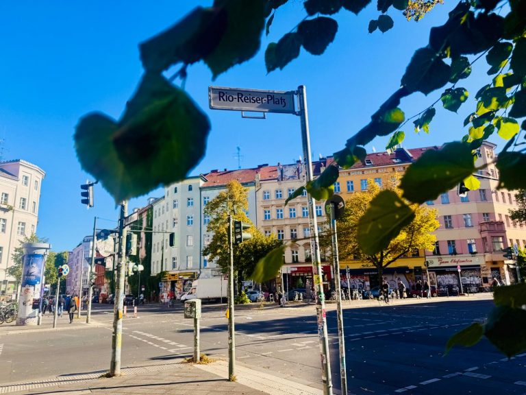 Straßenschild "Rio-Reiser-Platz" in Berlins pulsierendem Kreuzberg, mit bunten Gebäuden und Bäumen im Hintergrund unter strahlend blauem Himmel - perfekt für eine kulinarische Stadtführung durch diesen lebendigen Kiez.
