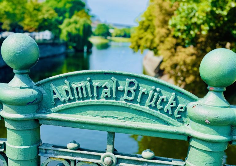 Grünes Metallschild mit der Aufschrift "Admiral-Brücke" auf einer Brücke über einen ruhigen Fluss mit Bäumen im Hintergrund, eine bezaubernde Szene, die oft während einer kulinarischen Stadtführung in Berlin erkundet wird.