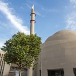 Moderne Moschee mit hohem Minarett, abgerundeter Kuppel, Baum im Vordergrund und blauem Himmel mit Wolken.