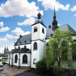 Historisches Kirchengebäude mit zwei Türmen und einem Baum unter einem blauen Himmel mit Wolken. In der Nähe parken Autos.