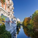 Ein von Herbstbäumen und klassischen Gebäuden gesäumter Kanal, der eine malerische Kulisse bildet, in der charmante Cafés unter einem klaren blauen Himmel köstliche Spezialitäten aus der Küche servieren.