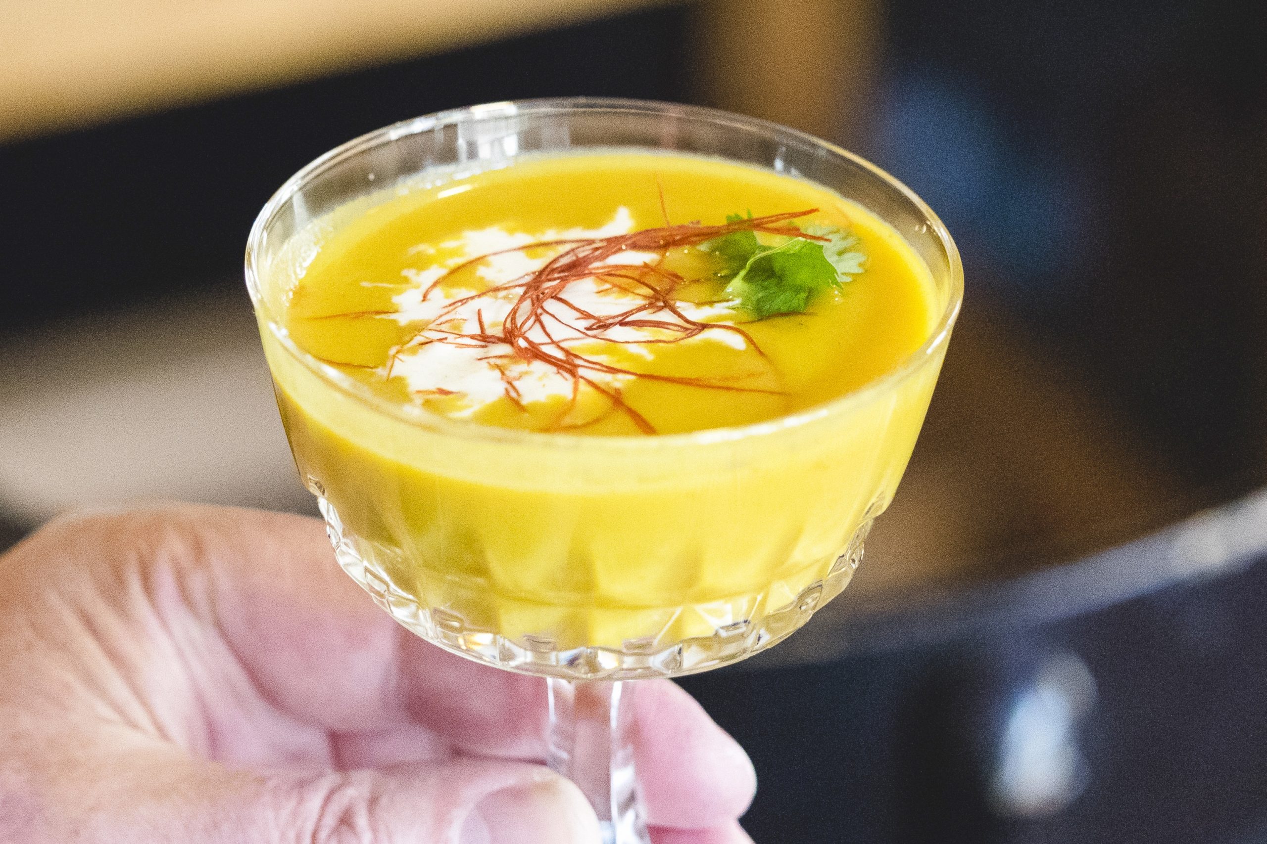 Eine Hand hält ein Glas voller cremiger gelber Suppe, elegant garniert mit Sahne und einem Zweig Koriander – ein perfekter Start für jede Junggesellenabschiedsfeier.