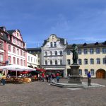 Historischer europäischer Platz mit farbenfrohen Gebäuden, einer Statue, Straßencafés und Menschen, die auf einer Kopfsteinpflasterstraße spazieren.