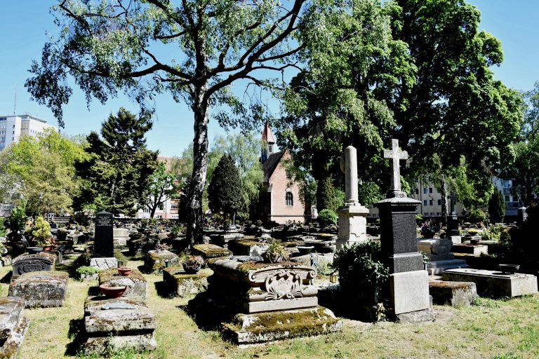 Alter Friedhof mit Grabsteinen und Bäumen in Nürnberg-Gostenhof, im Hintergrund eine Backsteinkirche und städtische Gebäude - perfekt, um sie bei einer kulinarischen Stadtführung zu erkunden.