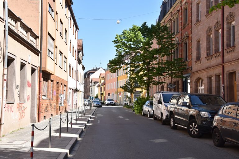 Eine sonnige Straße in Gostenhof, Nürnberg, gesäumt von geparkten Autos und bunten Häusern - perfekt für eine kulinarische Stadtführung unter Bäumen und einem klaren blauen Himmel.