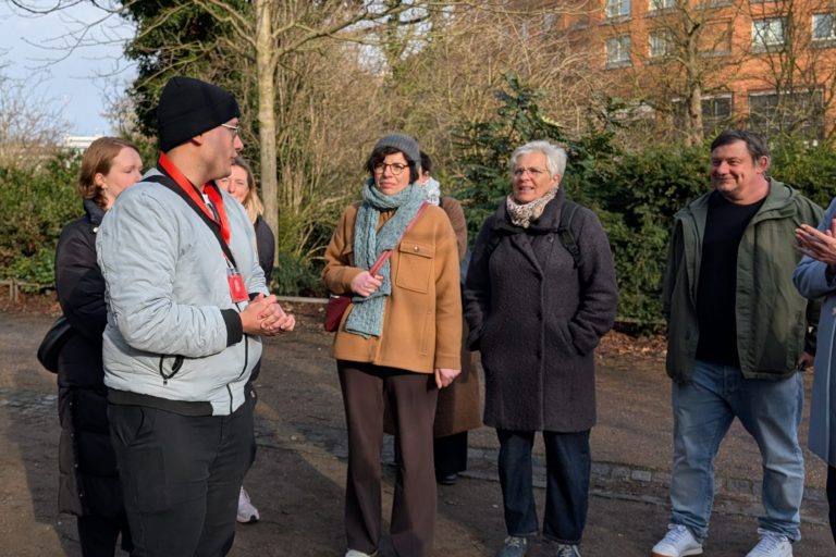 Eine Gruppe von Menschen genießt eine kulinarische Stadtführung, während sie an einem sonnigen Tag im Freien, umgeben von Bäumen und Grün, in der Bremer Altstadt einem Führer zuhört.
