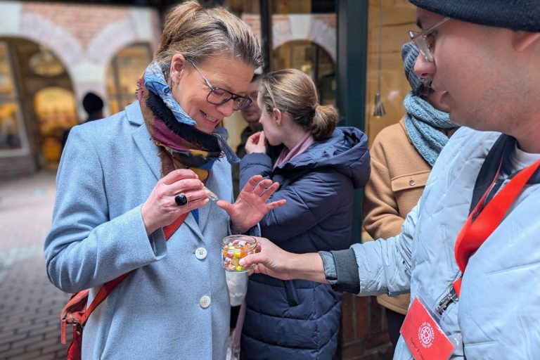 Eine lächelnde Frau nimmt während der Kulinarischen Stadtführung in der Bremer Altstadt einen kleinen Becher mit Süßigkeiten von einer Person entgegen, die ein rotes Schlüsselband trägt.