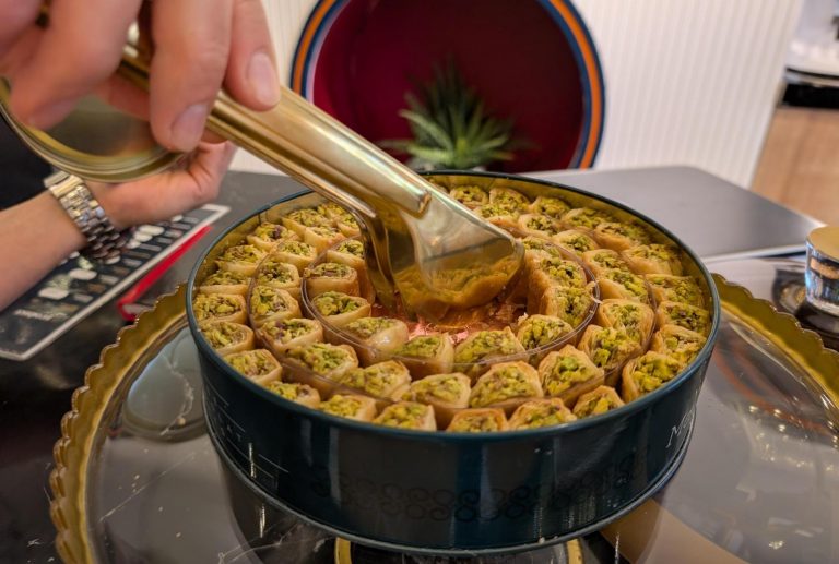 Eine Hand nimmt mit einer Zange ein Stück mit Pistazien gefülltes Baklava aus einer runden Dose, das während einer kulinarischen Stadtführung in Heidelbergs charmanter Altstadt genossen werden soll.