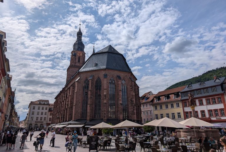 Cafétische im Freien und Menschen vor einer großen, historischen Kirche in der Heidelberger Altstadt – der perfekte Rahmen für einen kulinarischen Stadtrundgang über diesen bezaubernden europäischen Stadtplatz.