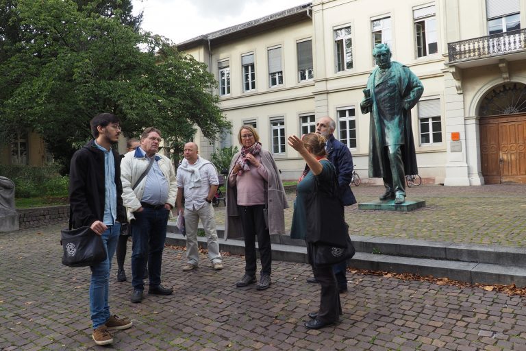Eine Gruppe von Menschen steht in der Nähe einer Statue vor einem großen Gebäude in der Heidelberger Altstadt, während eine Frau gestikuliert und spricht – vielleicht führt sie die Gruppe gerade auf einem kulinarischen Stadtrundgang durch die historische Innenstadt.