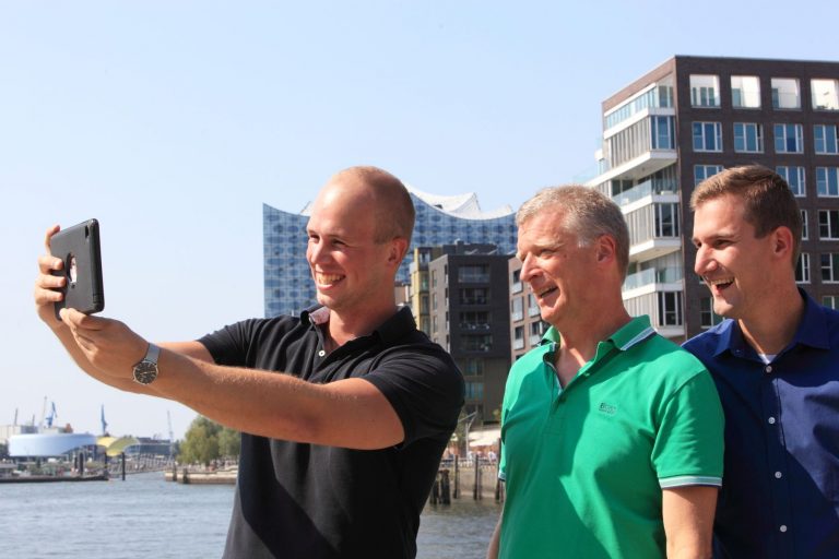 Drei Männer machen ein Selfie am Wasser in der HafenCity, mit der atemberaubenden modernen Skyline von Hamburg als Hintergrund.