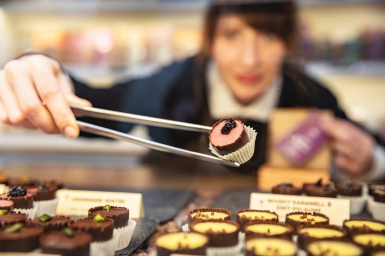 Eine Frau nimmt während einer kulinarischen Stadtführung in der Mannheimer Neckarstadt mit einer Zange ein Schokoladendessert aus einer Auslage mit verschiedenen Pralinen auf.