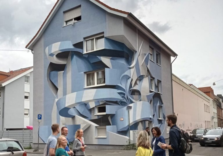 Eine Gruppe von Menschen steht vor einem Gebäude in der Mannheimer Neckarstadt, dessen Fassade mit großer, blauer, abstrakter 3D-Straßenkunst verziert ist.