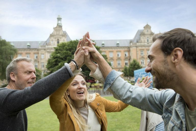 Eine Gruppe von vier Personen in einem Park, die mit einem gemeinsamen High-Five vor der historischen Kulisse Hamburgs feiern, schafft eine energiegeladene Szene, die an eine temperamentvolle Stadtrallye erinnert.