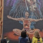 Im Mannheimer Jungbusch-Quadrat wird ein großes Wandbild betrachtet, das einen meditativen Menschen mit freiliegender Anatomie und aus dem Körper verzweigten Wurzeln zeigt.