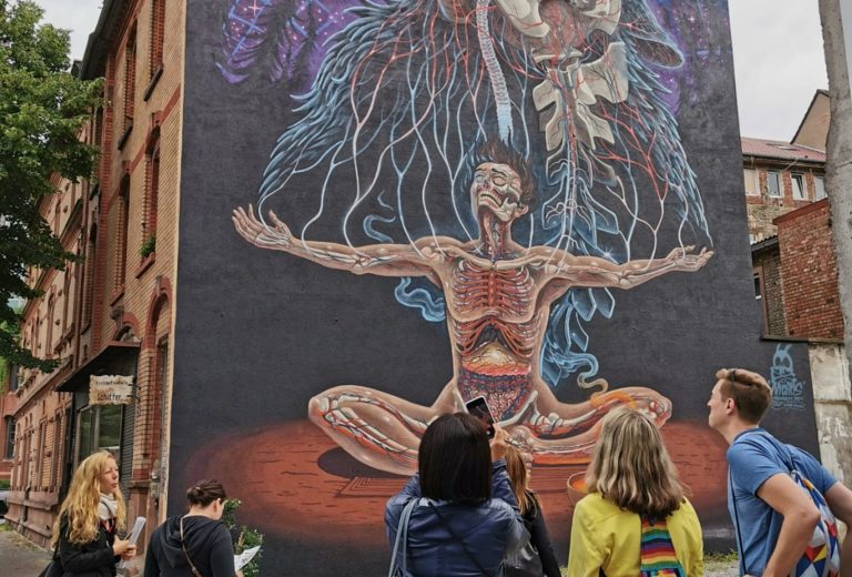 Im Mannheimer Jungbusch-Quadrat wird ein großes Wandbild betrachtet, das einen meditativen Menschen mit freiliegender Anatomie und aus dem Körper verzweigten Wurzeln zeigt.