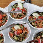Sechs Schalen frischer Salat mit Quinoa, Tomaten, Gurken und Radieschen, jeweils mit einem Löffel, auf einem weißen Tablett - perfekt zum Genießen nach einer kulinarischen Stadtführung Mannheim durch den lebendigen Mannheimer Jungbusch oder der Erkundung des berühmten Mannheimer Quadrats.