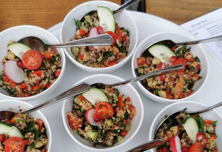 Sechs Schalen frischer Salat mit Quinoa, Tomaten, Gurken und Radieschen, jeweils mit einem Löffel, auf einem weißen Tablett - perfekt zum Genießen nach einer kulinarischen Stadtführung Mannheim durch den lebendigen Mannheimer Jungbusch oder der Erkundung des berühmten Mannheimer Quadrats.