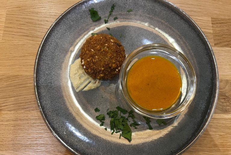 Ein Falafelbällchen mit Soße, Dip und gehackten Kräutern auf einem grauen Teller auf einem Holztisch - perfekt zum Probieren bei einer Kulinarischen Stadtführung Mannheim durch den lebendigen Jungbusch oder das historische Quadrate.