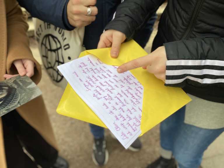 Eine Person zeigt auf einen handgeschriebenen Brief in einem gelben Umschlag für die Stadtrallye München.