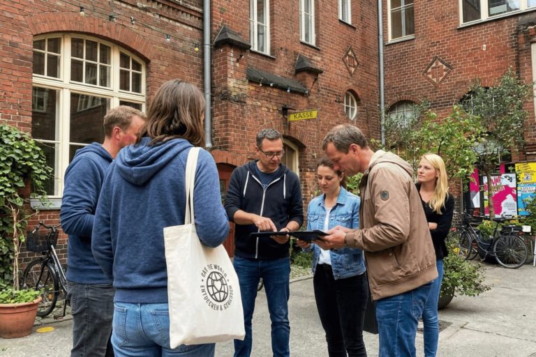 Eine Gruppe von Menschen steht vor einem Backsteingebäude in Berlin-Mitte und schaut sich während eines Outdoor-Escape-Games gemeinsam Unterlagen oder eine Karte an.