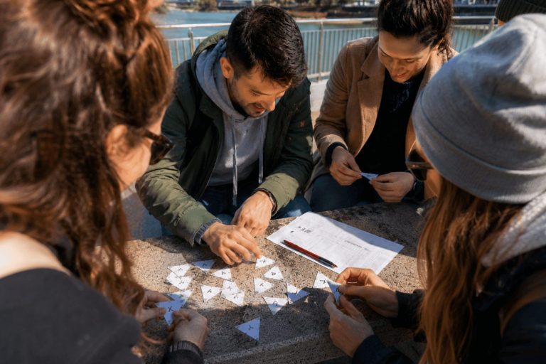 Vier Personen sitzen draußen in Carlstadt bei Düsseldorf um einen Tisch herum und lösen gemeinsam im Rahmen eines Outdoor-Escape-Games ein Dreieck-Rätsel, während Papier und Stift griffbereit liegen.