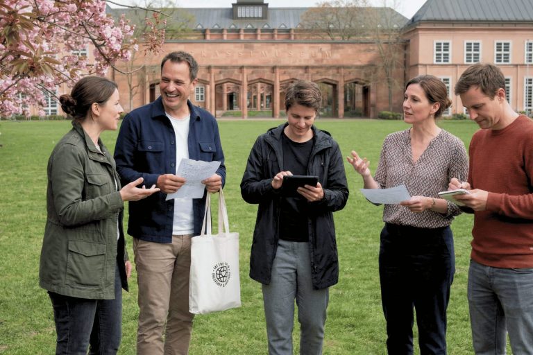 Fünf Personen stehen draußen auf einer Wiese im Graphischen Viertel in Leipzig, unterhalten sich und lächeln, während sie Zettel und ein Tablet in den Händen halten und sich auf ein Outdoor-Escape-Game in der Nähe historischer Gebäude vorbereiten.