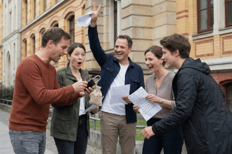 Fünf Menschen im Graphischen Viertel in Leipzig feiern begeistert im Freien, schauen auf ein Handy und halten bedrucktes Karopapier in den Händen – sie genießen ein Outdoor-Escape-Game-Abenteuer.