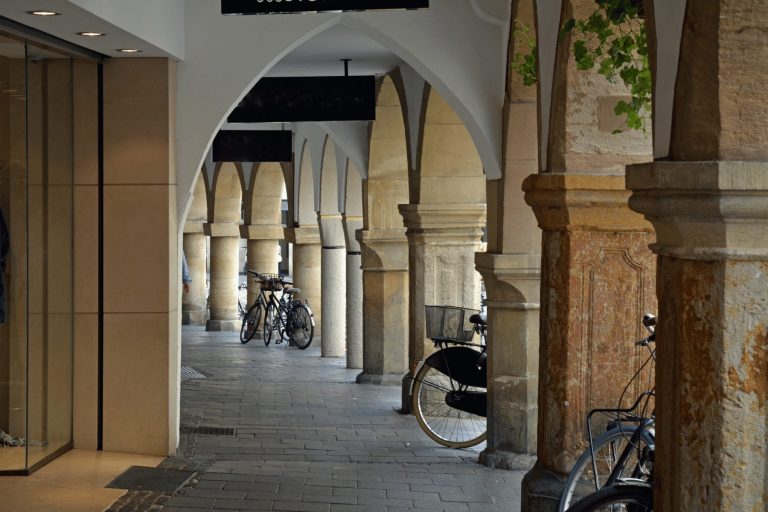 Ein Steinbogen mit geparkten Fahrrädern entlang eines überdachten Gehwegs in Münsters charmanter Altstadt – der perfekte Ort, um Ihr nächstes „Outdoor Escape Game“-Abenteuer zu beginnen.