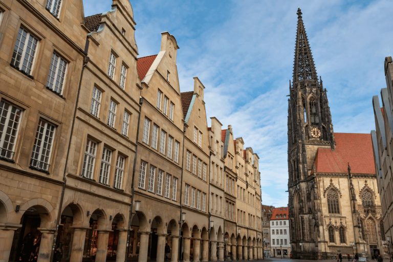 Historische Gebäude mit gewölbten Arkaden in der Münsters Altstadt stehen neben einem hohen gotischen Kirchturm, alles unter blauem Himmel.