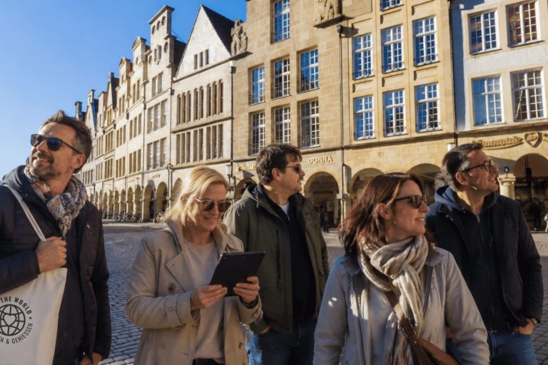 Fünf lächelnde Menschen stehen auf einem sonnigen Kopfsteinpflasterplatz und sind bereit, ein Outdoor-Escape-Game in der Münsters Altstadt zu beginnen, im Hintergrund sind historische Gebäude zu sehen.