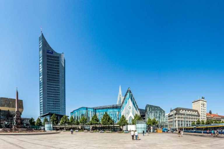 Panoramablick auf moderne Gebäude, darunter ein hoher Turm und Glaskonstruktionen, unter einem klaren blauen Himmel.