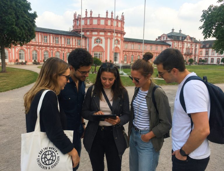 Fünf Personen stehen vor einem großen, prunkvollen Gebäude in Wiesbaden und lächeln gemeinsam, während sie bei einem Outdoor-Escape-Game auf ein Tablet blicken.