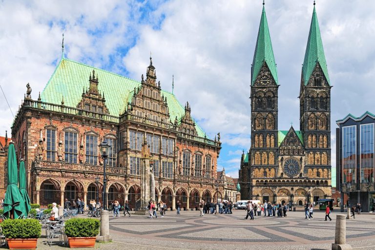 Ein historischer Platz in der Bremer Altstadt mit kunstvoll verzierten Gebäuden und Kirchtürmen mit zwei Türmchen unter einem teilweise bewölkten Himmel – perfekt für ein „Outdoor Escape Game“-Abenteuer.