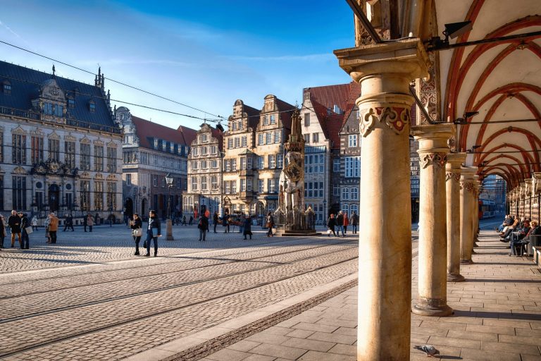 Unter blauem Himmel schlendern Menschen über den historischen Altstädter Platz in Bremen, umgeben von kunstvoll verzierten Gebäuden und gewölbten Säulen – eine ideale Kulisse für ein Outdoor-Escape-Game.