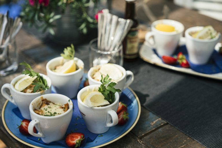 Tassen Suppe garniert mit Kräutern und Zitronenscheiben auf einem blauen Tablett mit Erdbeeren, auf einem rustikalen Holztisch.