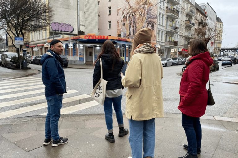 Vier Personen stehen an einer Straßenecke im historischen Zentrum von Hamburg, an einem bewölkten Tag mit Jacken bekleidet – vielleicht bereiten sie sich gerade auf ein „Outdoor Escape Game“-Abenteuer vor.