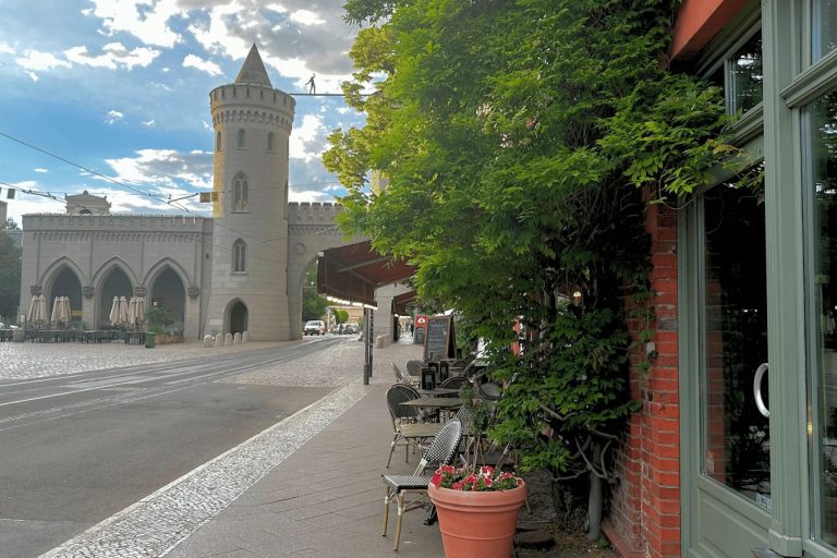 Café-Sitzplätze im Freien an einer Kopfsteinpflasterstraße im Potsdamer Zentrum, mit einem historischen Steintor und einem Turm im Hintergrund – ideal zum Entspannen nach einem Outdoor-Escape-Game oder Ihrem letzten Outdoor-Abenteuer.