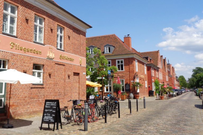 Historische rote Backsteinstraße im Sommer mit Cafés, Fahrrädern und Bäumen unter einem blauen Himmel.
