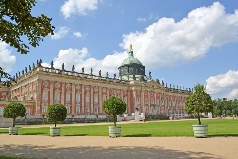 Historischer Palast mit verzierter Fassade, Kuppel und Statuen unter einem blauen Himmel mit flauschigen Wolken. Umgeben von gepflegtem Rasen.