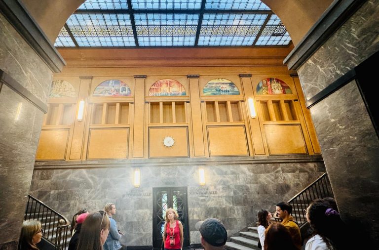 In einem großen Saal mit Wandmalereien, Marmorwänden und einer gläsernen Decke lauscht eine Gruppe während einer kulinarischen Stadtführung in Berlin einem Führer.