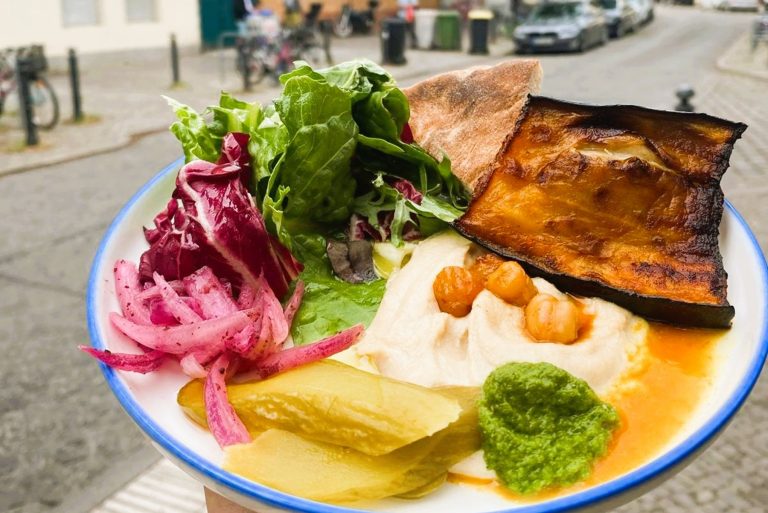 Ein Teller mit Hummus, gegrillten Auberginen, Grünzeug, Essiggurken, rosa Zwiebeln und grüner Soße - perfekt für eine kulinarische Stadtführung in Berlin Neukölln, die im Freien stattfindet.