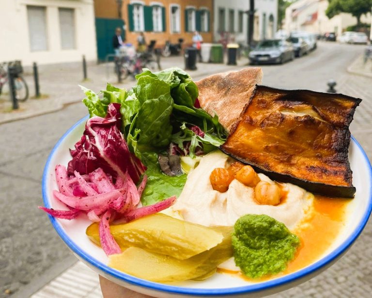Ein bunter Teller mit Hummus, Salat, Essiggurken, gegrilltem Gemüse und Brot auf einer Straße während einer kulinarisch veganen Stadtführung in Berlin Neukölln.