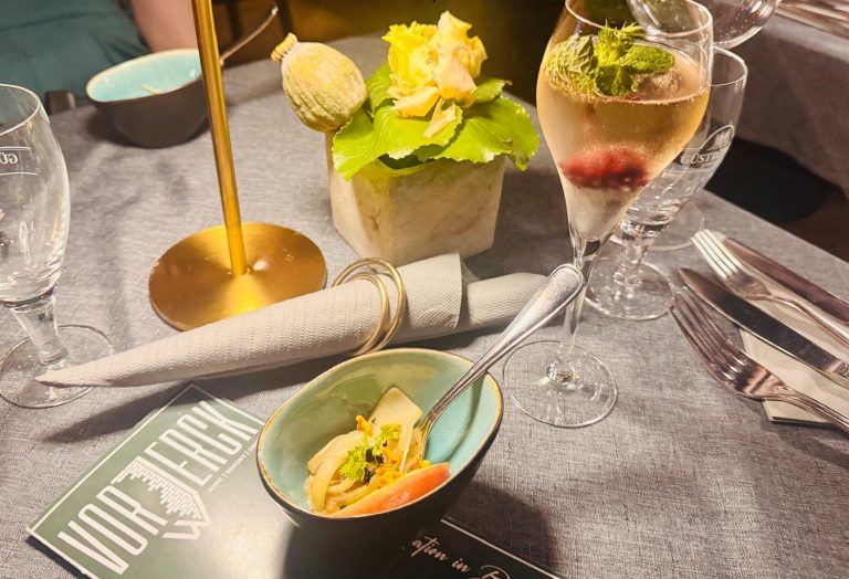 Auf einer kulinarischen Stadtführung in Berlin erwartet die Gäste ein gedeckter Tisch mit Blumenschmuck, Vorspeisenschale, gerollter Serviette und einem Glas prickelndem Getränk.