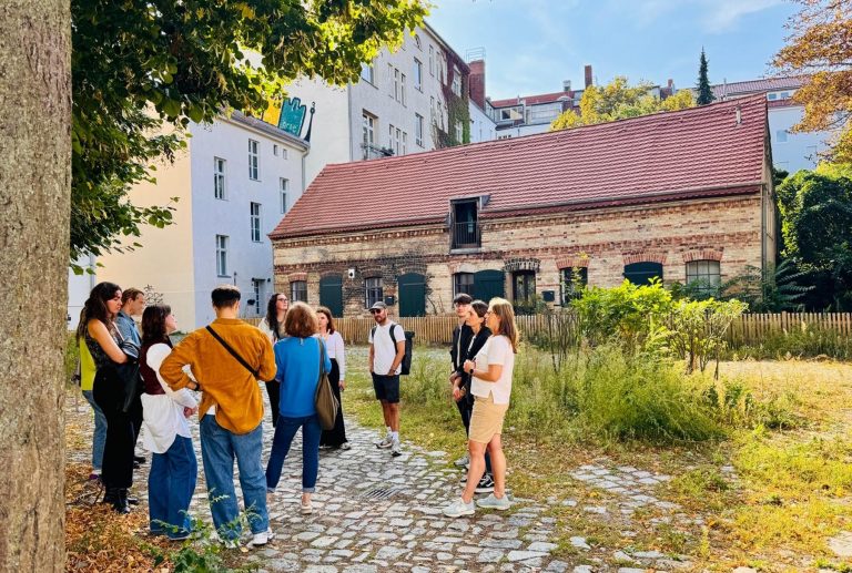 Eine Gruppe von Menschen steht an einem sonnigen Tag während einer kulinarischen Stadtführung in Berlin Neukölln auf einem Kopfsteinpflasterweg in der Nähe eines Backsteingebäudes und von Bäumen und unterhält sich.