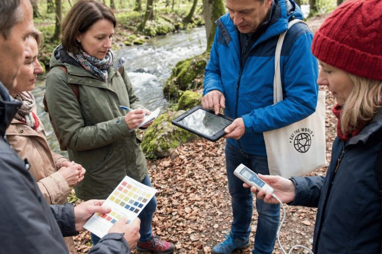 Fünf Personen betreiben wissenschaftliche Forschung an einem Bach in der Nähe der Residenz in München und nutzen dabei während eines Outdoor-Escape-Games ein Tablet, ein Notebook, eine Farbtabelle und ein Messgerät.