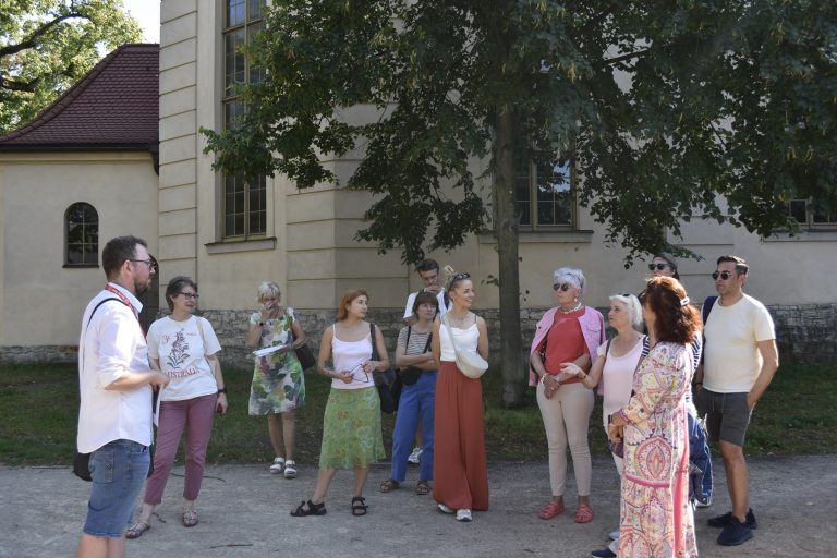 Eine Gruppe von Menschen hört einem Redner im Freien in der Nähe eines großen Gebäudes und Baums in Babelsberg aufmerksam zu, möglicherweise im Rahmen einer Kulinarischen Stadtführung, bei der die reiche Geschichte und die Aromen von Potsdam erkundet werden.
