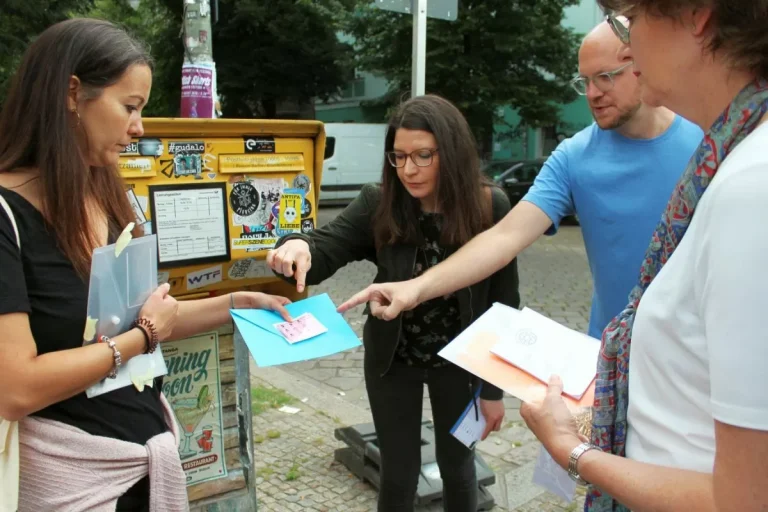 Vier Menschen diskutieren in ein Gespräch vertieft neben einem gelben Briefkasten Dokumente vor der Kulisse des pulsierenden Berlins. Vielleicht planen sie eine spannende Tour durch Schöneberg, dessen reiche Geschichte und lebendige Kultur im Mittelpunkt stehen.